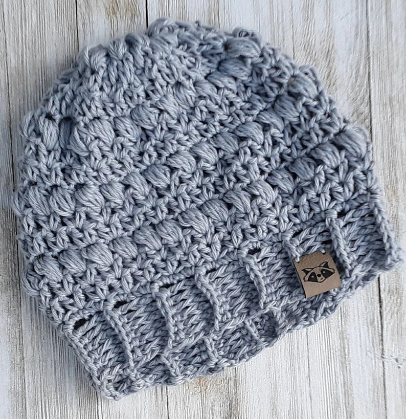Crocheted Dahoon Slouch Beanie in silver-gray cotton blend yarn, laid flat on a white wood surface, showing puff stitch texture and ribbed band with a small raccoon label
