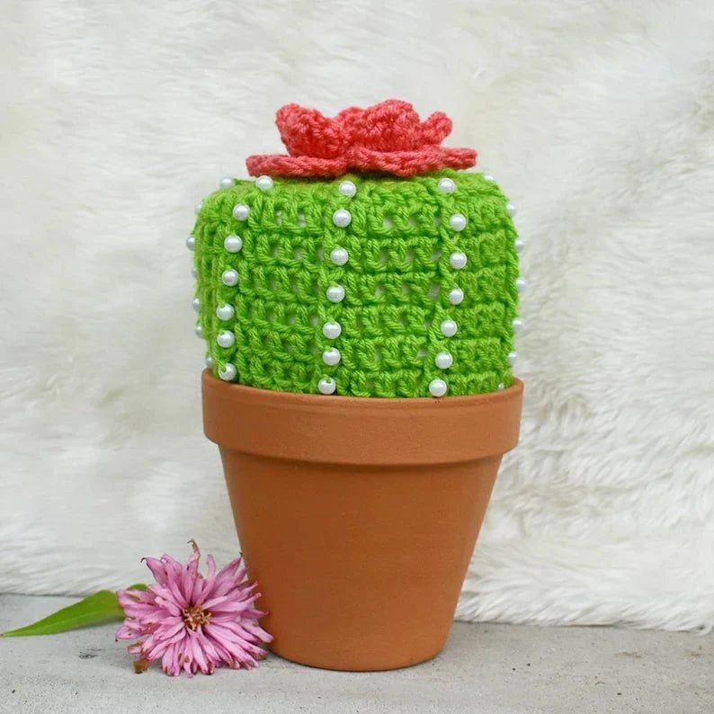 Crocheted cactus toilet paper cover with white pearl bead spines and pink flower topper, displayed in a terracotta pot with a pink daisy beside it