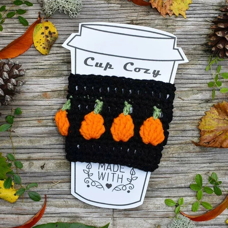 Black crochet pumpkin cup cozy on a paper cup card surrounded by autumn leaves, pine cones, and greenery on a rustic wood surface