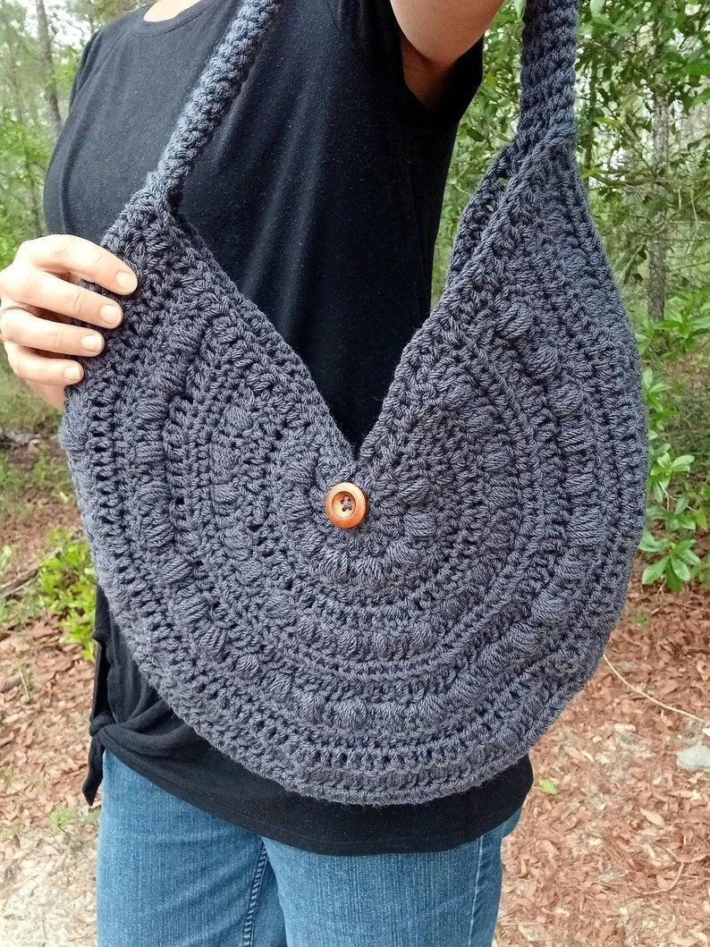 Person wearing a crescent-shaped crochet purse with puff stitch texture and wooden button closure, made from gray worsted weight cotton yarn