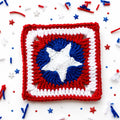 A crocheted square patch featuring a Captain America shield design with red, white, and blue yarn, surrounded by festive red, white, and blue confetti and star sprinkles on a white background.