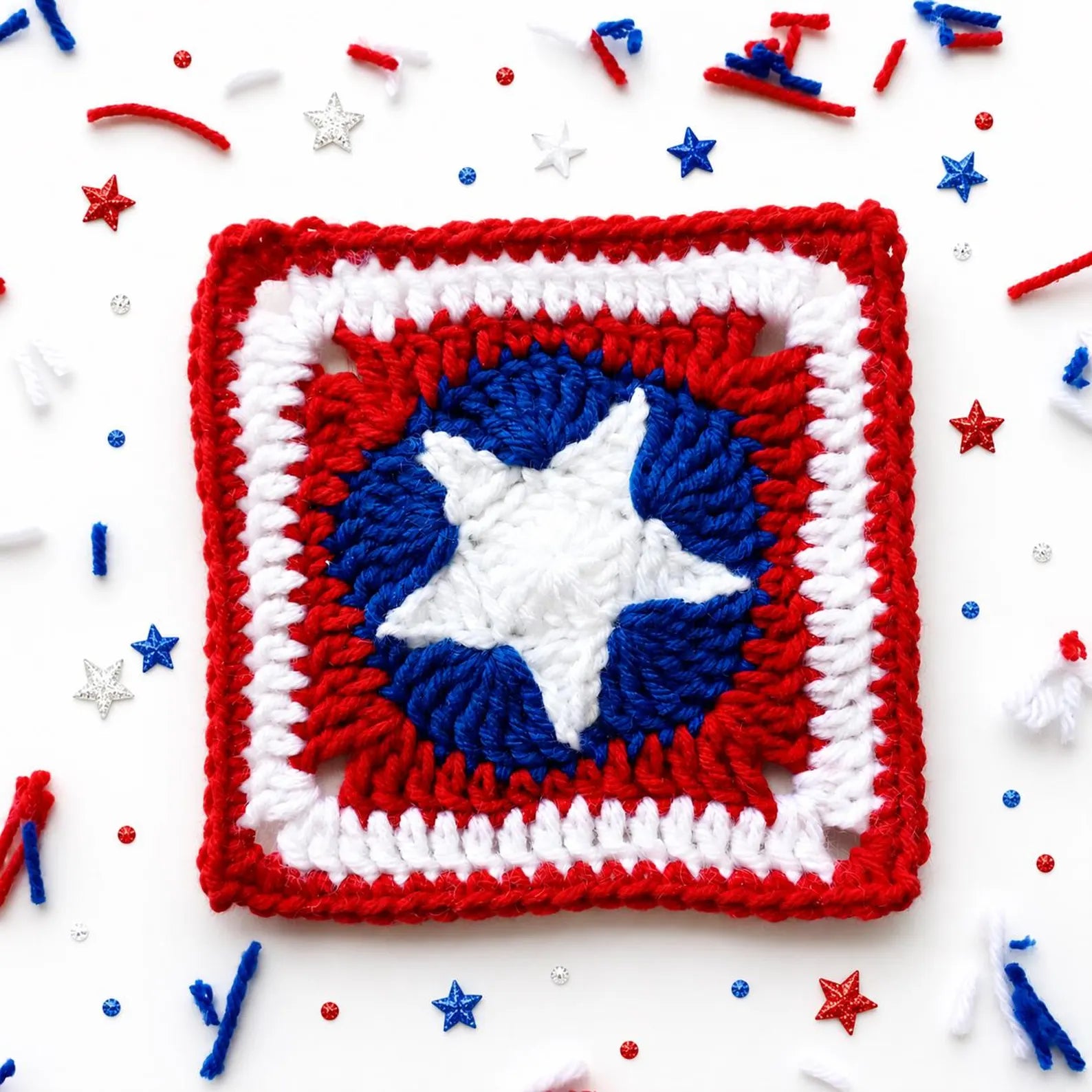 A crocheted square patch featuring a Captain America shield design with red, white, and blue yarn, surrounded by festive red, white, and blue confetti and star sprinkles on a white background.