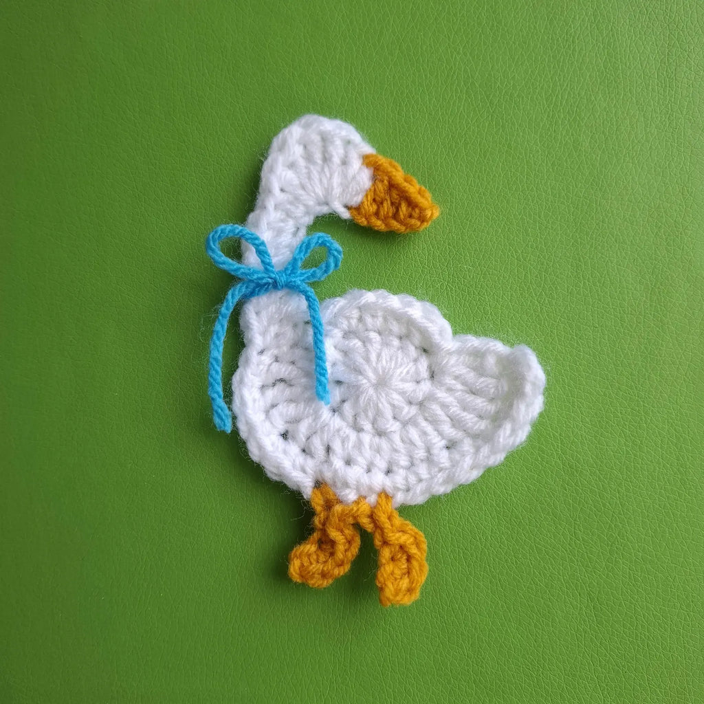 Crocheted white goose applique with blue ribbon bow and yellow feet on green