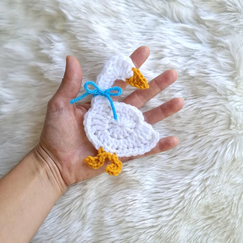 Crocheted white goose applique with blue bow and yellow feet in hand