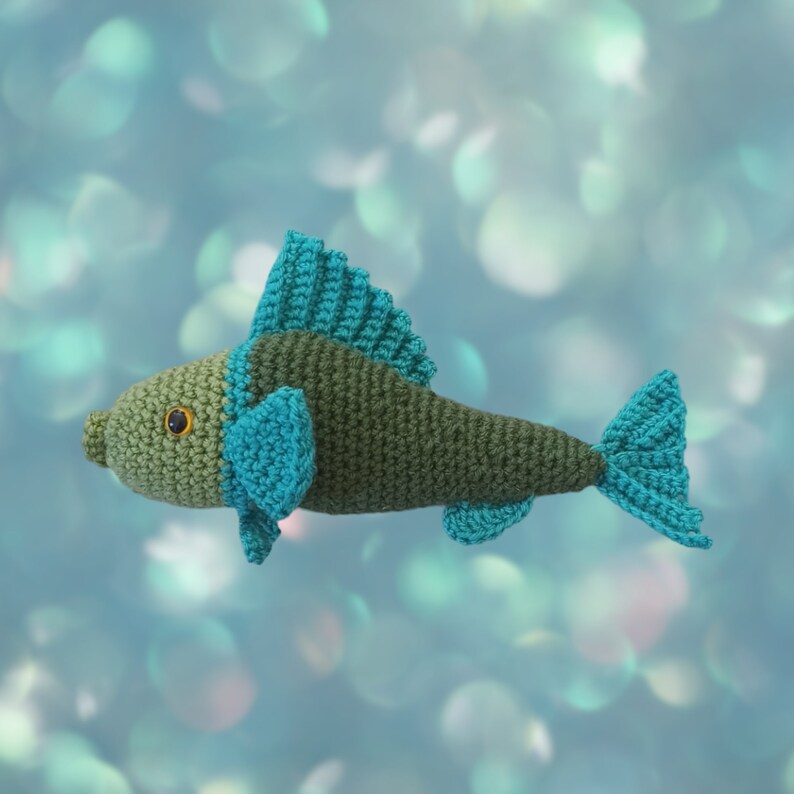 A crocheted fish toy with a dark olive green body and bright turquoise-blue fins, gills, and tail, shown against a soft, out-of-focus blue and teal bokeh background.