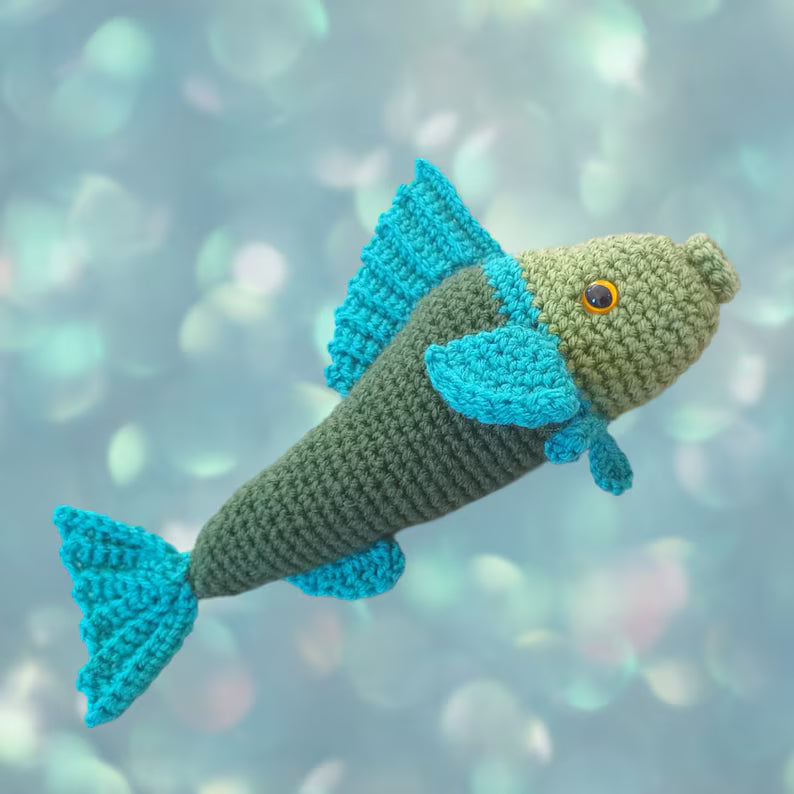 A crocheted fish toy with a dark green body and bright turquoise-blue fins, set against a soft, out-of-focus blue and teal bokeh background.