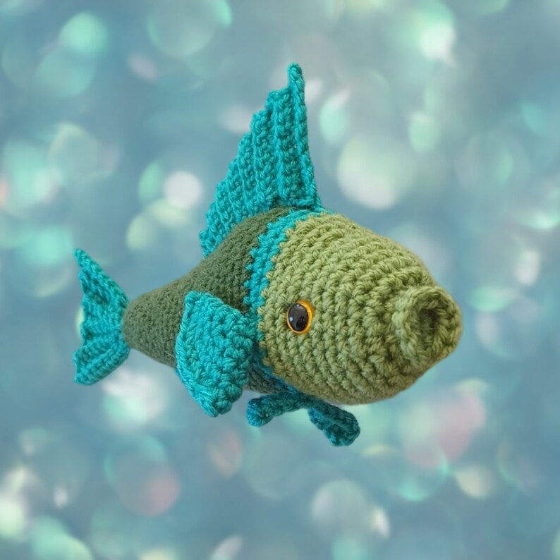 A crocheted fish toy with an olive green body, teal-blue fins and tail, and a small gold-and-black eye, shown against a soft blue bokeh background.