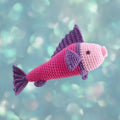A crocheted fish toy with a pink body, purple fin and tail, and a light pink face, shown against a soft blue-green bokeh background.
