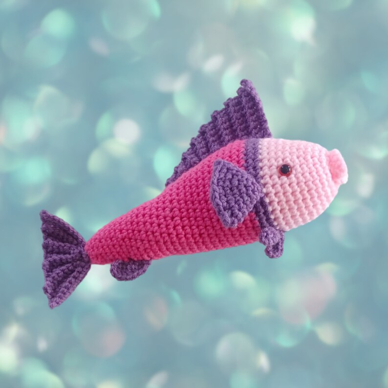 A crocheted fish toy with a pink body, purple fin and tail, and a light pink face, shown against a soft blue-green bokeh background.